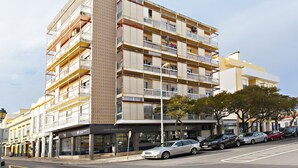 Un immeuble à plusieurs étages doté de balcons, avec un espace commercial au rez-de-chaussée et des voitures garées le long de la rue.