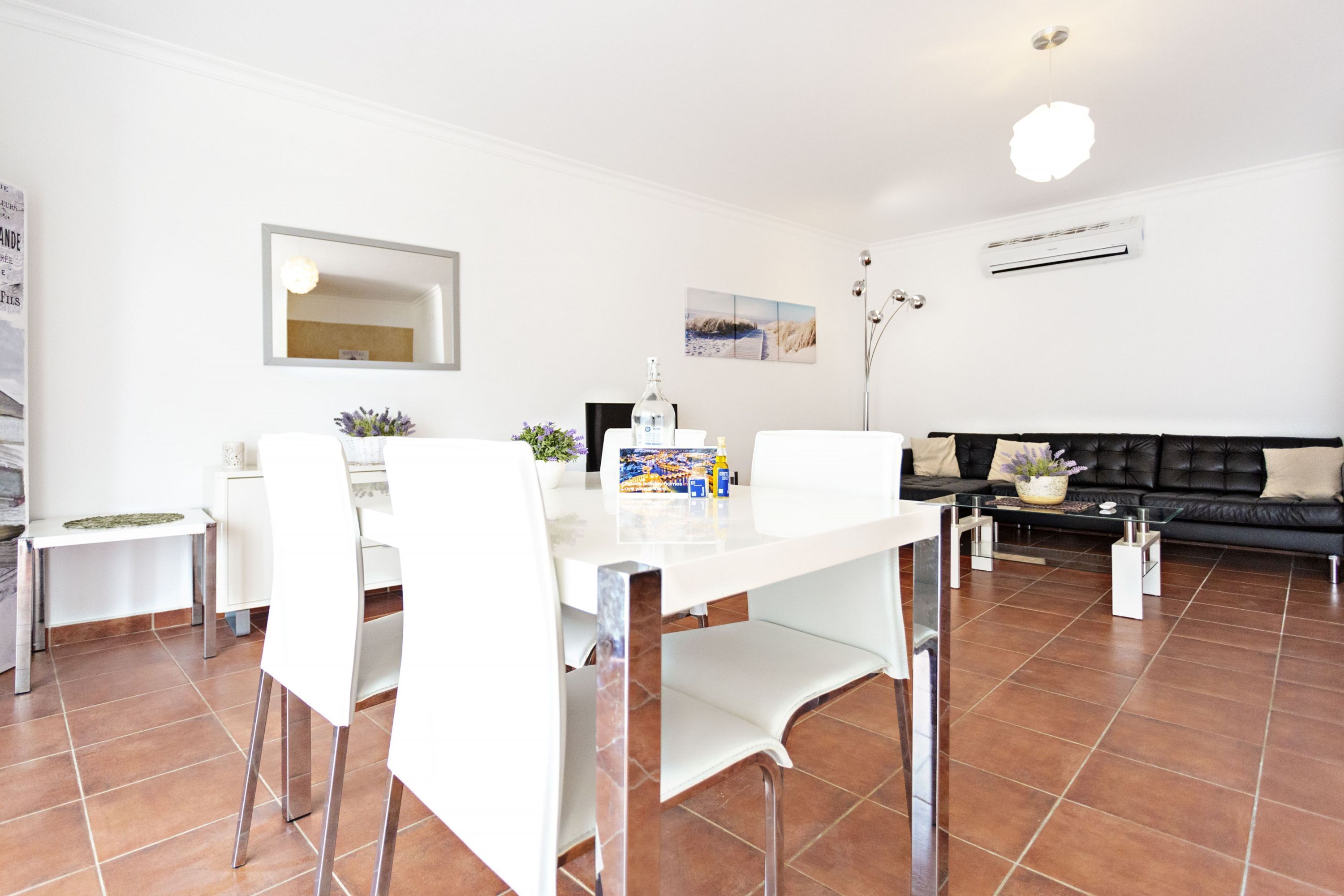 A modern living room with a dining area, featuring a white dining table, chairs, a black leather sofa, and a coffee table.