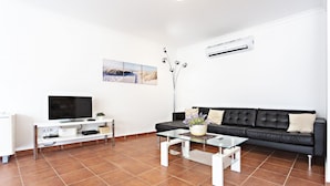 A modern living room with a black leather sofa, a glass coffee table, a white entertainment unit, and a large window. - Lavender in Tavira With 2 Bedrooms and 2 Bathrooms (Tavira)
