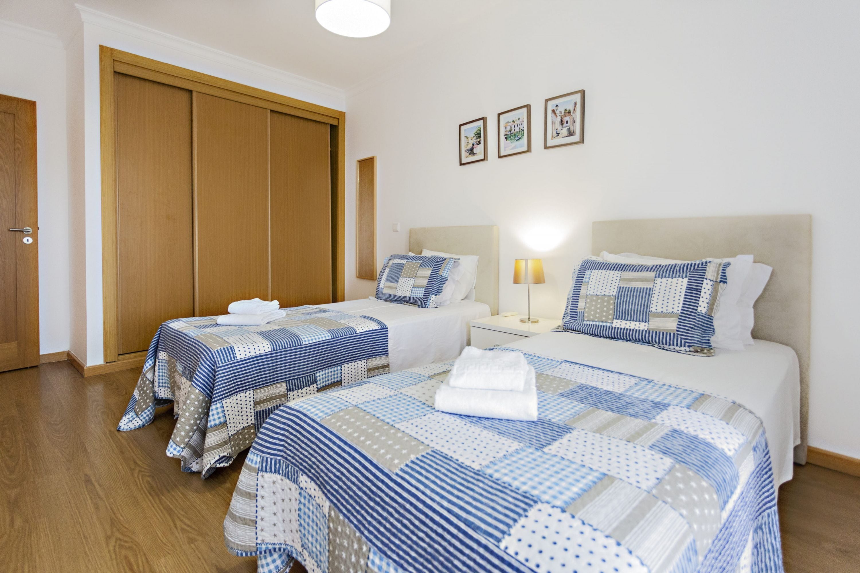 A hotel room with two beds, a wooden wardrobe, and a nightstand with a lamp.