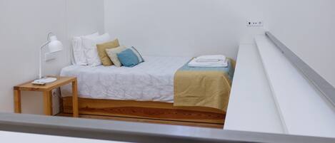A small, neatly arranged bedroom with a single bed, a bedside table with a lamp, and a power outlet on the wall.