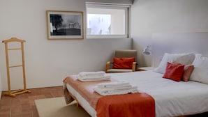 A neatly arranged bedroom with a bed, a chair, a ladder, a window, and a framed picture on the wall. - CV AI- Convento das Bernardas (Tavira)