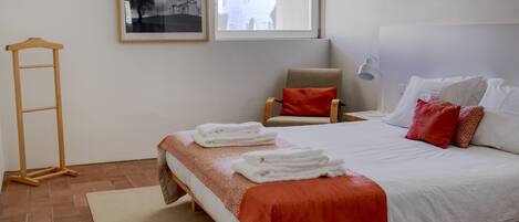 A neatly arranged bedroom with a bed, a chair, a ladder, a window, and a framed picture on the wall.