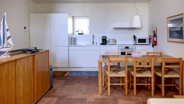 A compact kitchen with white cabinets, a dining table with wooden chairs, a small window, and a white pendant light. - CV AI- Convento das Bernardas (Tavira)