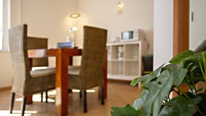 A dining area with a wooden table and wicker chairs, a white cabinet, and a potted plant. - QV 119 - Le Bleu Studio - Quinta Velha (Tavira)