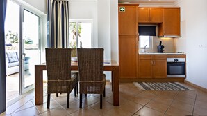 A dining area with wicker chairs, a wooden table, and a kitchen with wooden cabinets and a built-in oven. - QV 10 - Terrace of Peace Apartment - Quinta Velha (Tavira)