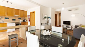 A modern kitchen with wooden cabinets, a dining area with a glass table, and a living room with a sofa and TV. - Cam lia in Tavira With 2 Bedrooms and 2 Bathrooms (Tavira)