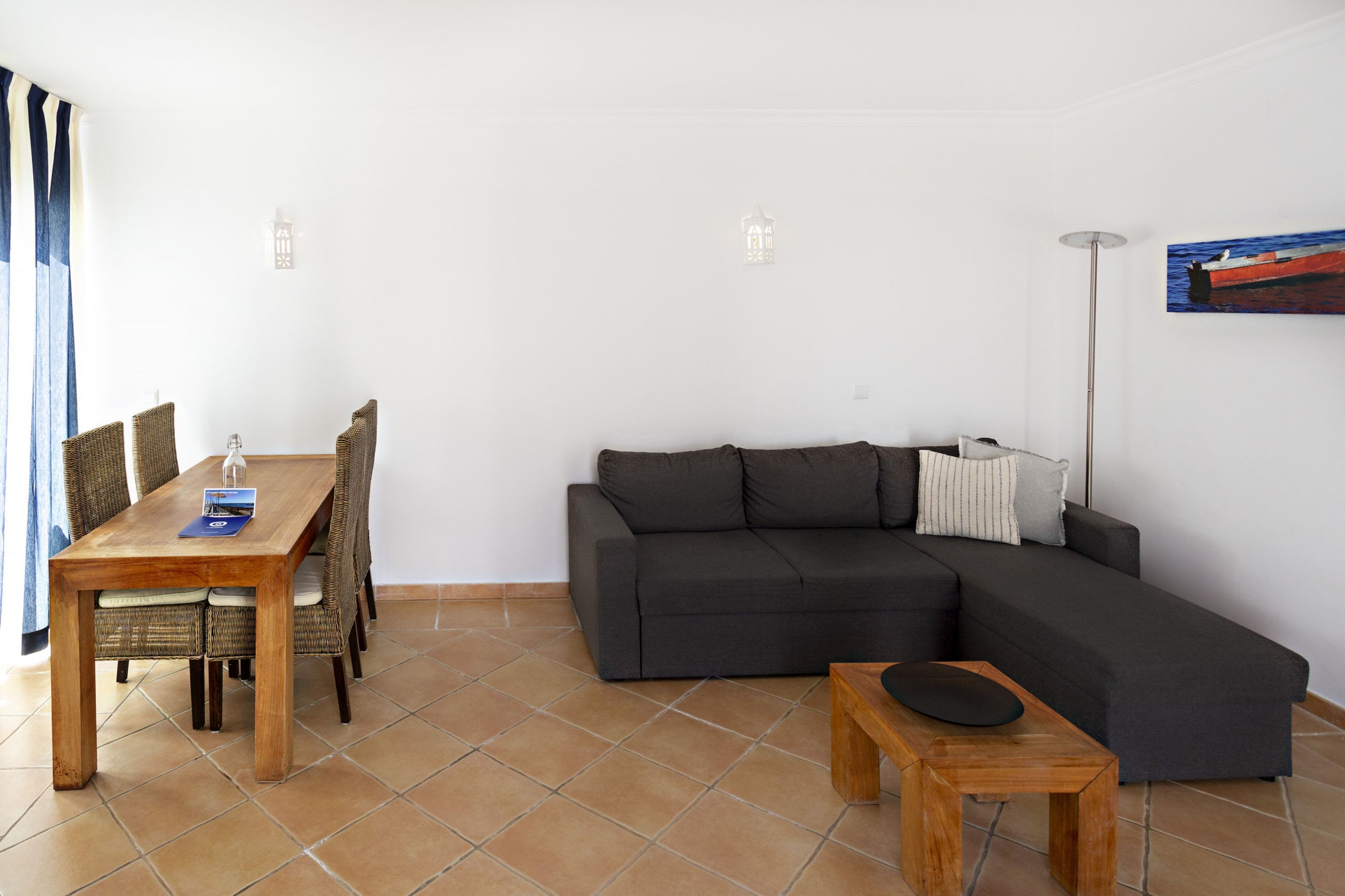 Ein Wohnzimmer mit einem dunkelgrauen Ecksofa, einem Holzesstisch mit Korbstühlen, einem kleinen Holznachttisch und einer Stehlampe.