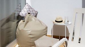 A room with a white chair, a beige bean bag chair, a small round table with a hat and a glass, and a window with a patterned pillow.