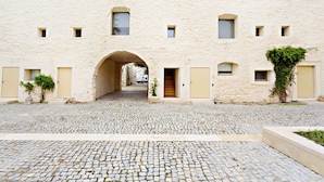 A cobblestone courtyard with a central archway leading to a building with multiple small windows and doors. - CV AR- Convento das Bernardas (Tavira)