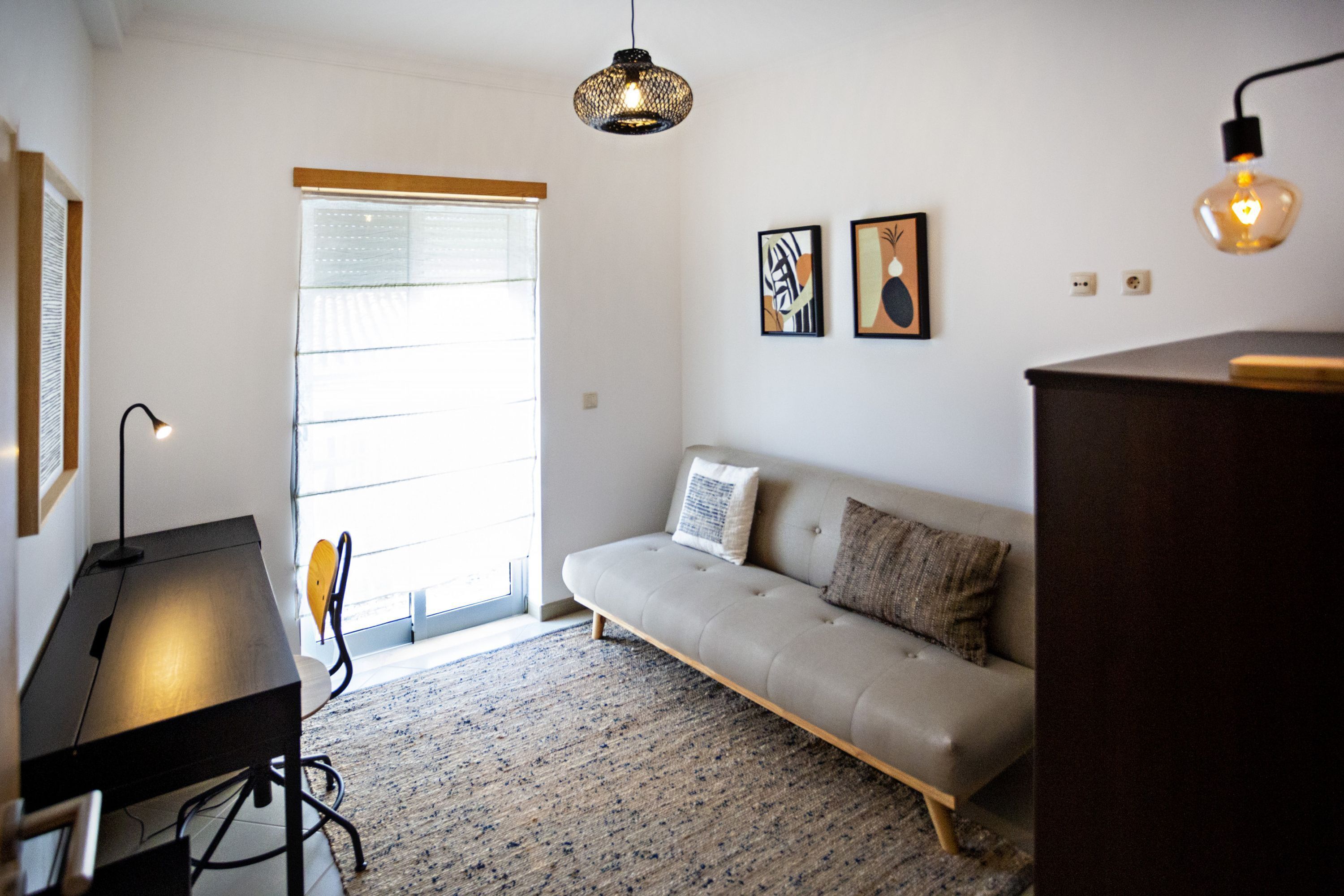 A modern living room with a sofa, a desk, and a lamp. There is a window with blinds and two wall-mounted artworks.
