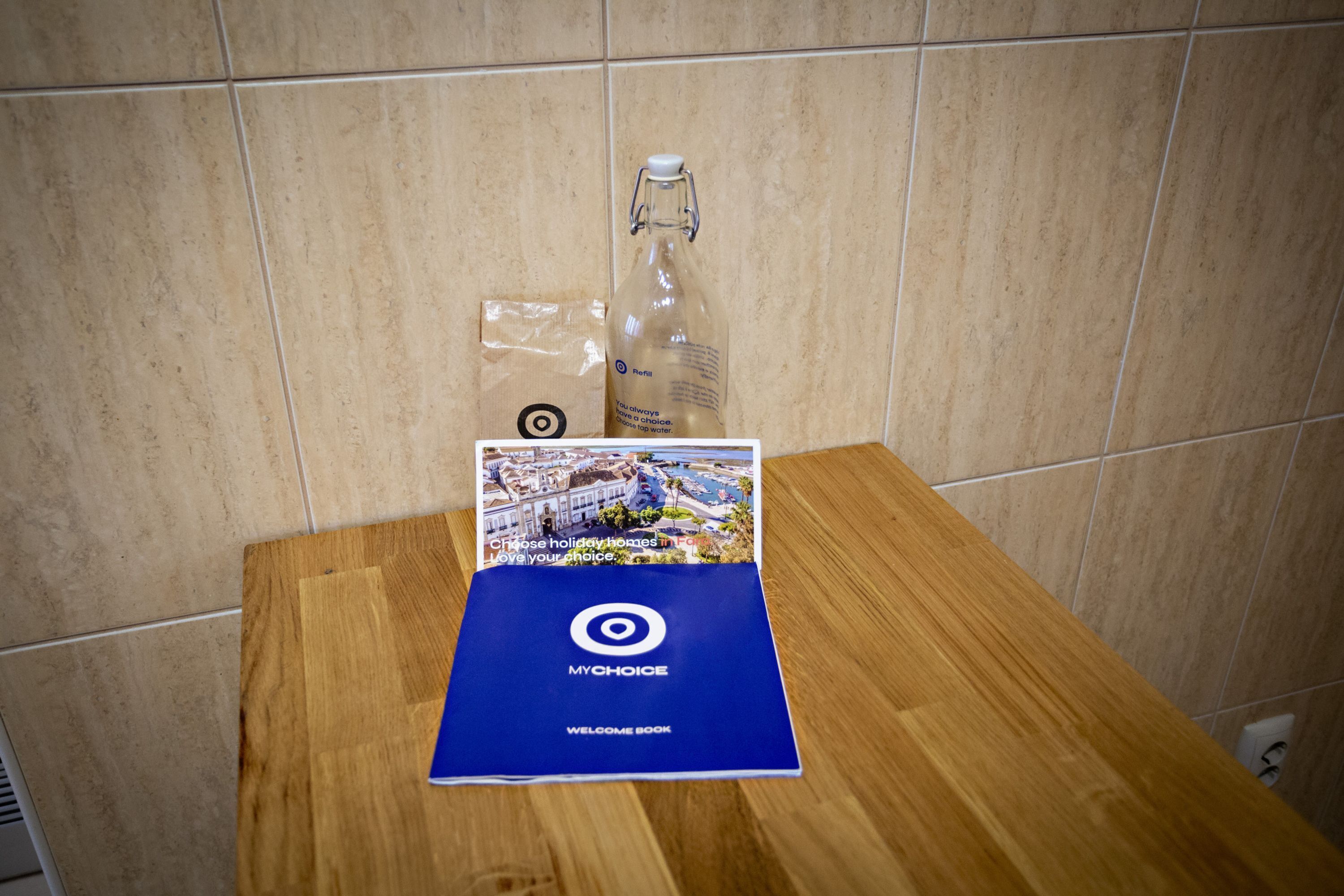 A welcome book and a bottle on a wooden table against a tiled wall.