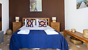 Un dormitorio con una cama grande, dos mesitas de noche con lámparas, un banco de madera y una estatua de Buda colgada en la pared.
