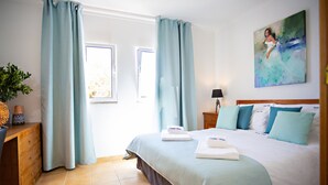 A hotel room with a bed, a nightstand, a lamp, a painting, and a window with curtains. - QV 21 - Straw Hat Apartment - Quinta Velha (Tavira)
