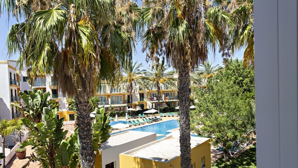 Een resort met een zwembad, palmbomen en een geel gebouw.