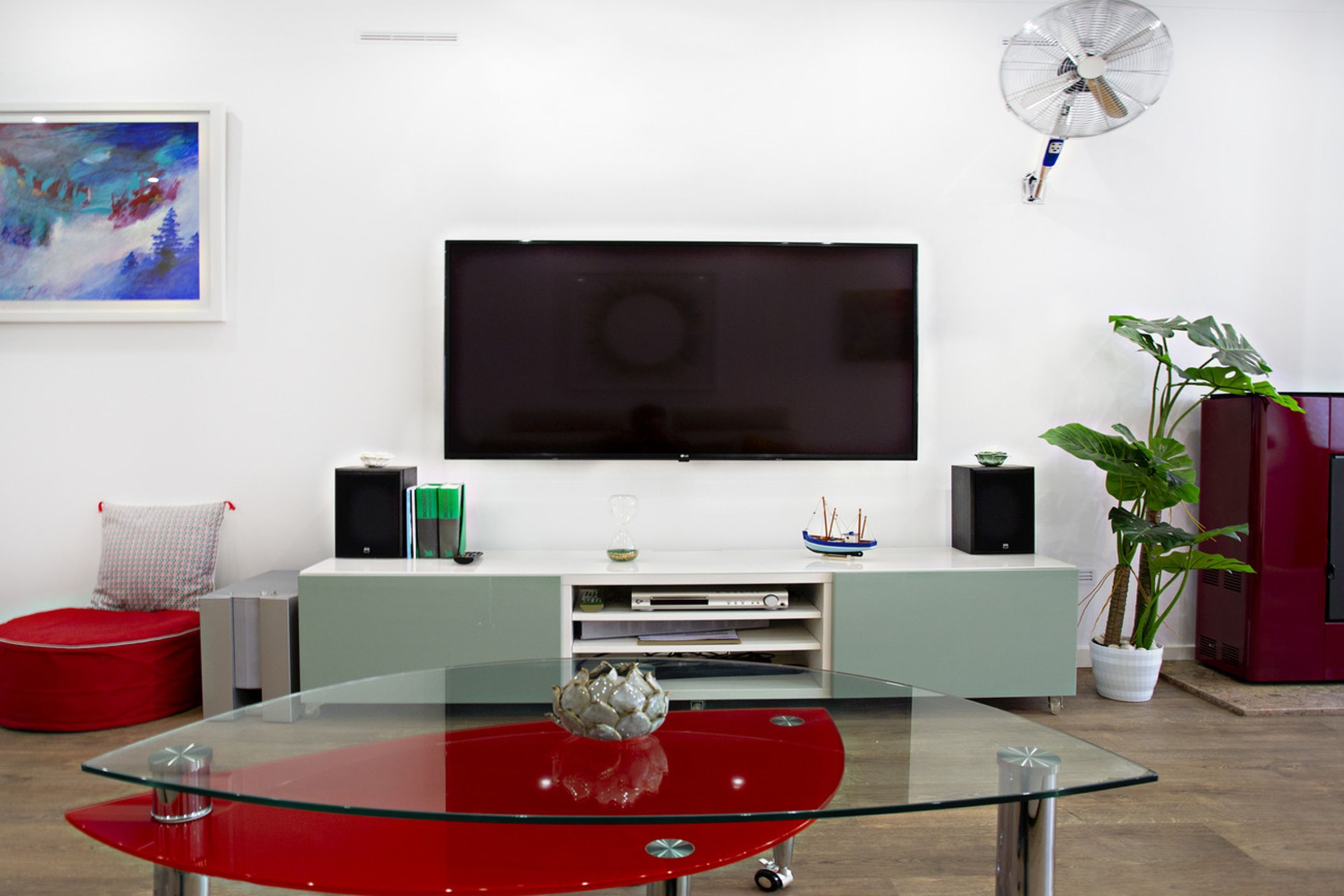 A modern living room with a glass-top coffee table, a flat-screen TV, a red bean bag, a potted plant, and a red cabinet.