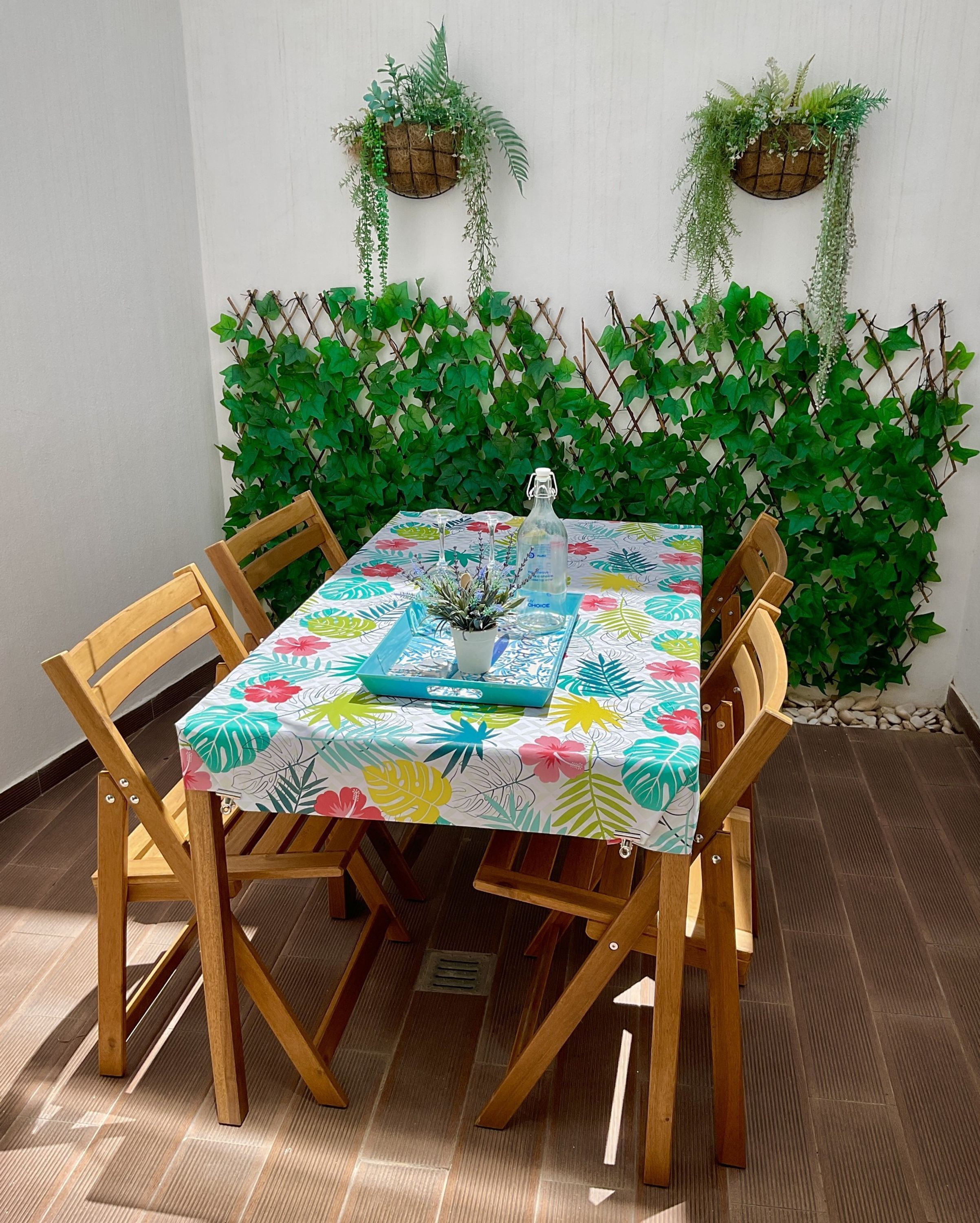 A table set with a floral tablecloth, surrounded by wooden chairs, against a backdrop of green foliage and a white wall.