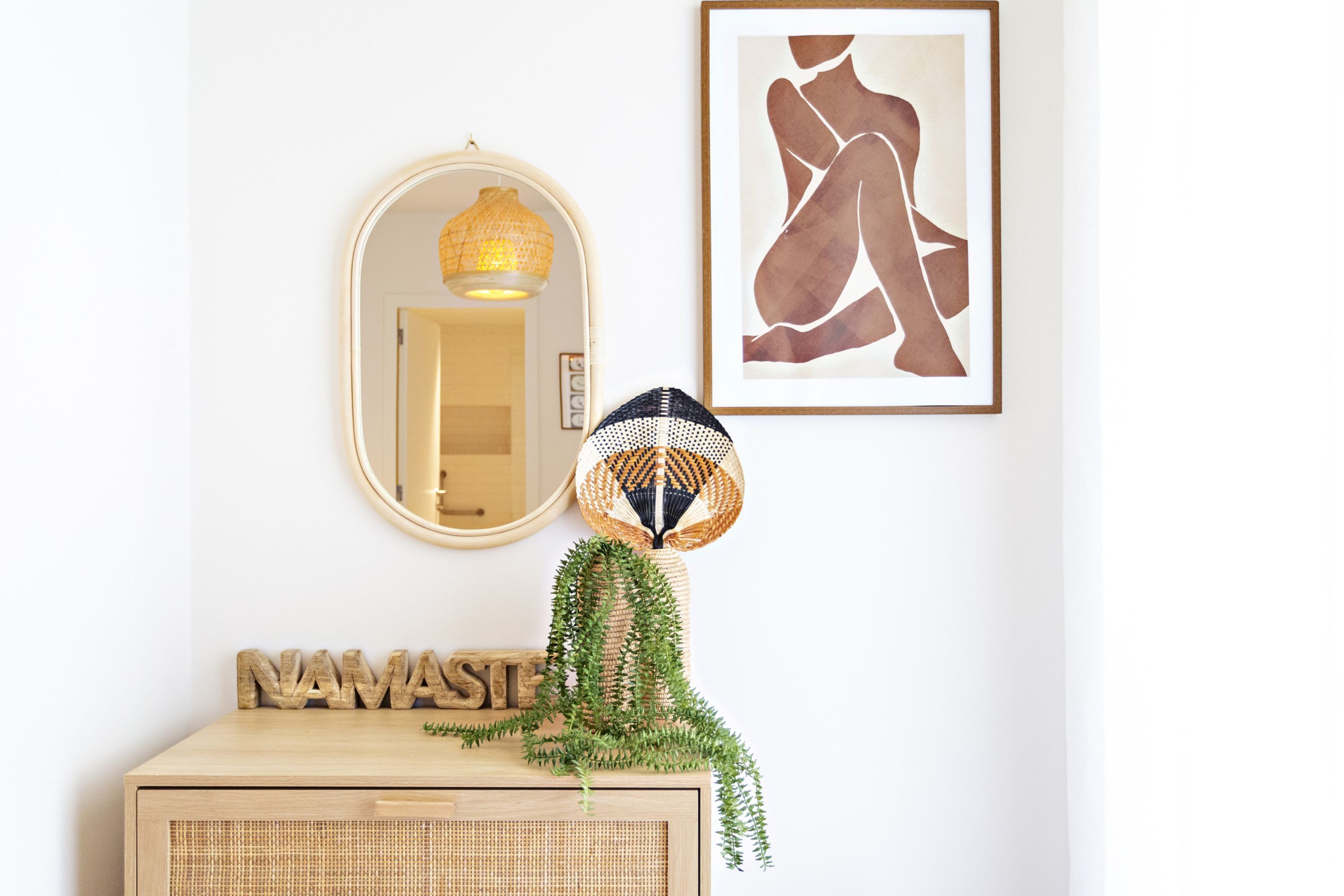 A wall-mounted mirror, a framed abstract artwork, a decorative lamp, and a wooden sign with the word 'NAMASTE' on a light-colored wooden dresser.