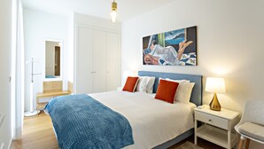 A bedroom with a large bed, a bedside table with a lamp, a painting on the wall, and a wooden bench in the corner. - Casas do Forte BE in Tavira (Tavira)
