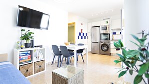 A modern living room with a flat-screen TV, a dining area with chairs, a kitchen with a fridge, oven, and washing machine. - Formosa Bay - Cabanas Bay (Tavira)