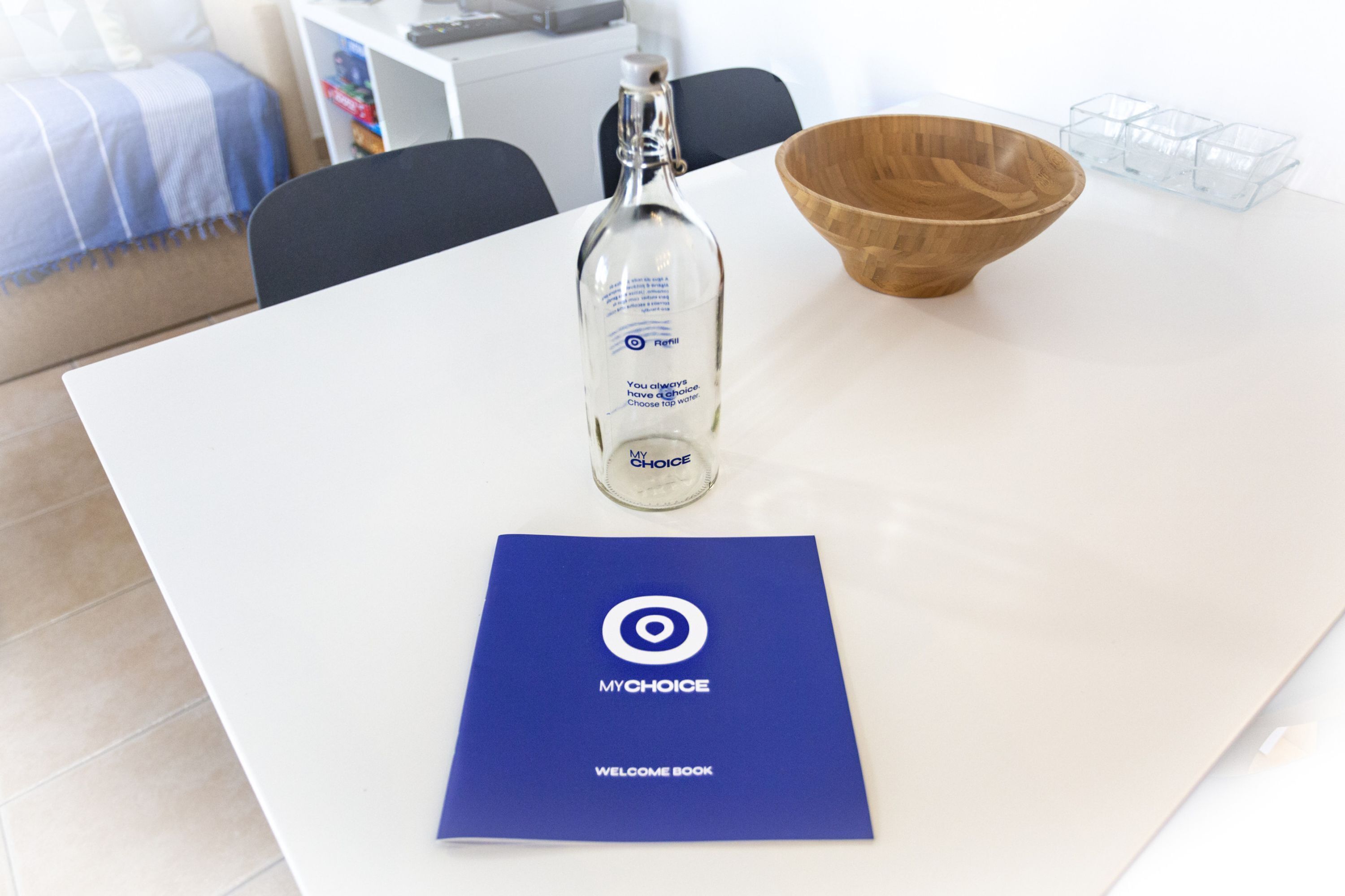 A white table with a clear glass bottle, a wooden bowl, and a blue welcome book.
