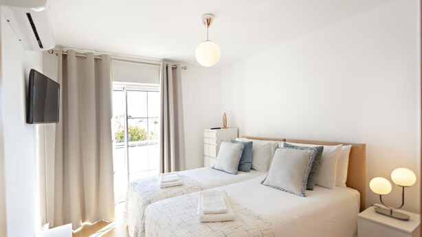 A modern bedroom with a large bed, bedside tables, a TV, and a window with curtains. - Formosa Bay - Cabanas Bay (Tavira)
