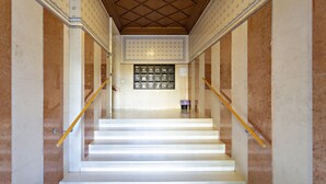 Una scala con pareti in marmo e soffitto decorato, che conduce a un pianerottolo con una vetrina di oggetti incorniciati.