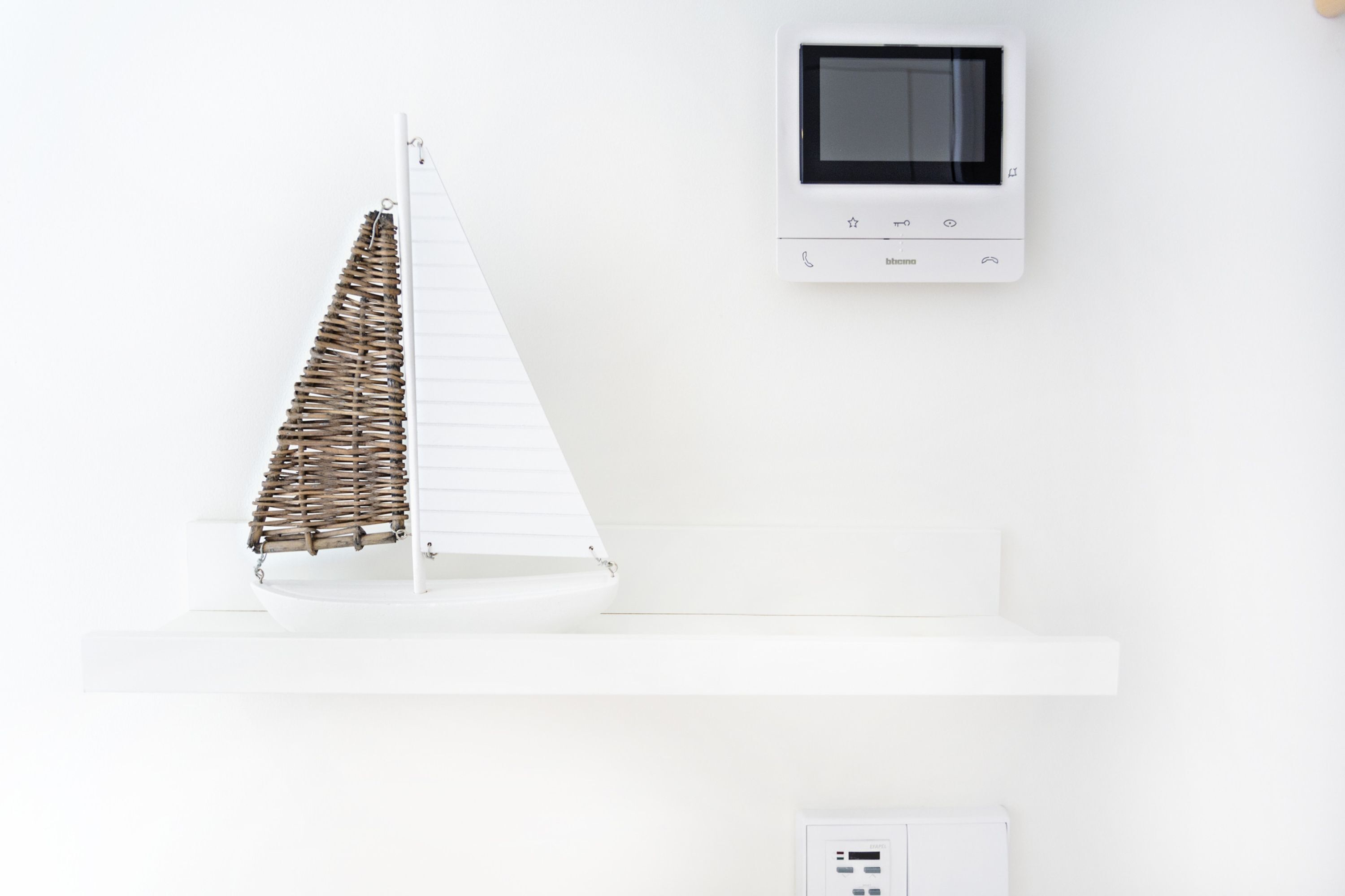 A white room with a model sailboat on a shelf, a thermostat, and a wall-mounted control panel.
