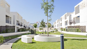 Un complexe résidentiel moderne, avec des bâtiments blancs, des jardins aménagés et un parterre circulaire central agrémenté d’un arbre.