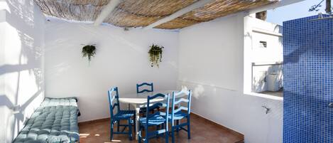 Un pequeño patio con una mesa y sillas, un banco acolchado y un techo de paja.