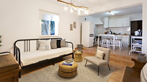 Un soggiorno moderno con divano, poltrona e pouf, angolo cottura con mobili bianchi e zona pranzo con sgabelli.