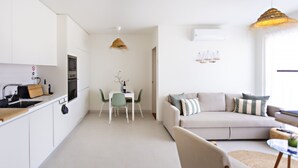 A modern kitchen with white cabinets, a dining table with chairs, and a living area with a sofa and cushions. - Casas do Forte EF - Casa de Cabanas (Tavira)