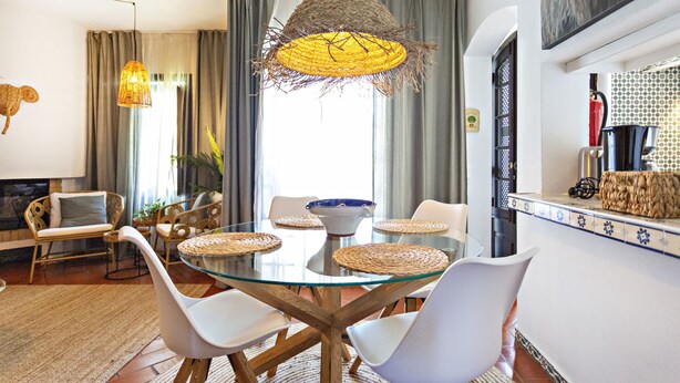 A dining area with a glass table, white chairs, woven placemats, and a hanging light fixture. - QV 14B - Casa da Pintora - Quinta Velha (Tavira)
