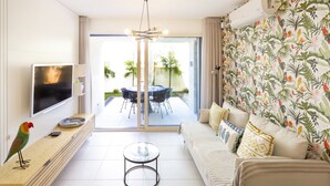A modern living room with a flat-screen TV, a wooden console table, a glass coffee table, a sofa with patterned cushions, and a large sliding glass door leading to an outdoor patio with a dining set.