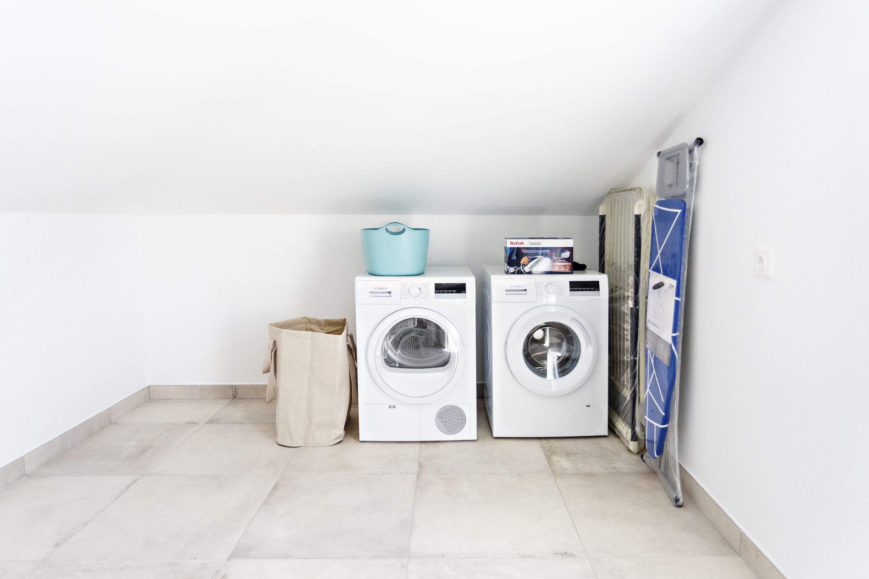 A laundry room with two white washing machines, a blue laundry basket, and a beige trash bag.