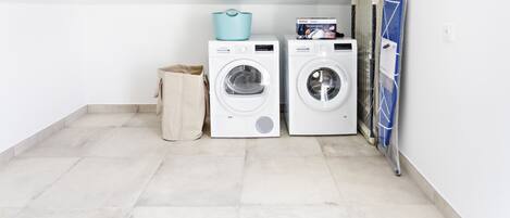 Une buanderie avec deux machines à laver blanches, un panier à linge bleu et un sac poubelle beige.