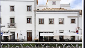 A white building with multiple windows and a tiled roof. There are outdoor tables and chairs with umbrellas, and a metal railing in the foreground. - Alagoa Garden View in Tavira (Tavira)