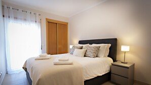 Una cama bien hecha con sábanas y almohadas blancas, una mesita de noche con lámpara y un armario de madera.