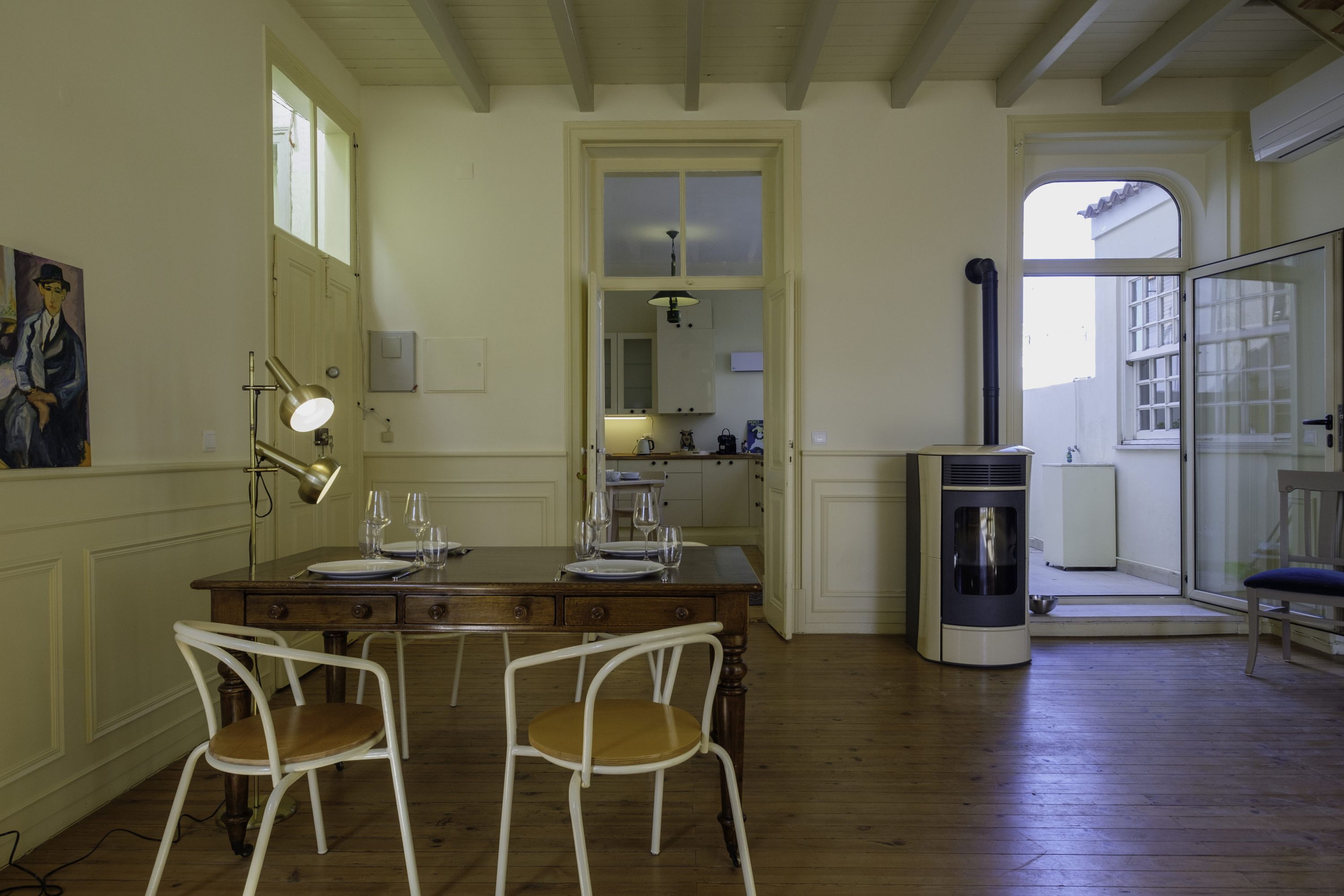 A dining area with a wooden table, two white chairs, a painting on the wall, and a kitchen visible through an open door.
