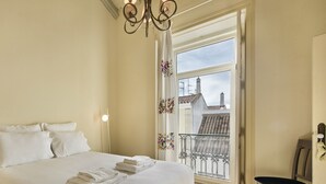 A hotel room with a large bed, a chandelier, a balcony with a view of buildings, and a chair. - Casa do Jornal in Vila Real de Santo Ant nio (Vila Real Santo Antonio)