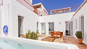 Un espace piscine avec une piscine blanche, des chaises en bois et une table. On y trouve également des plantes en pot et une rambarde rouge.