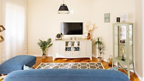 A living room with a TV, white cabinet, blue sofa, patterned rug, and decorative items. - Blue Peacock in Faro (Faro)