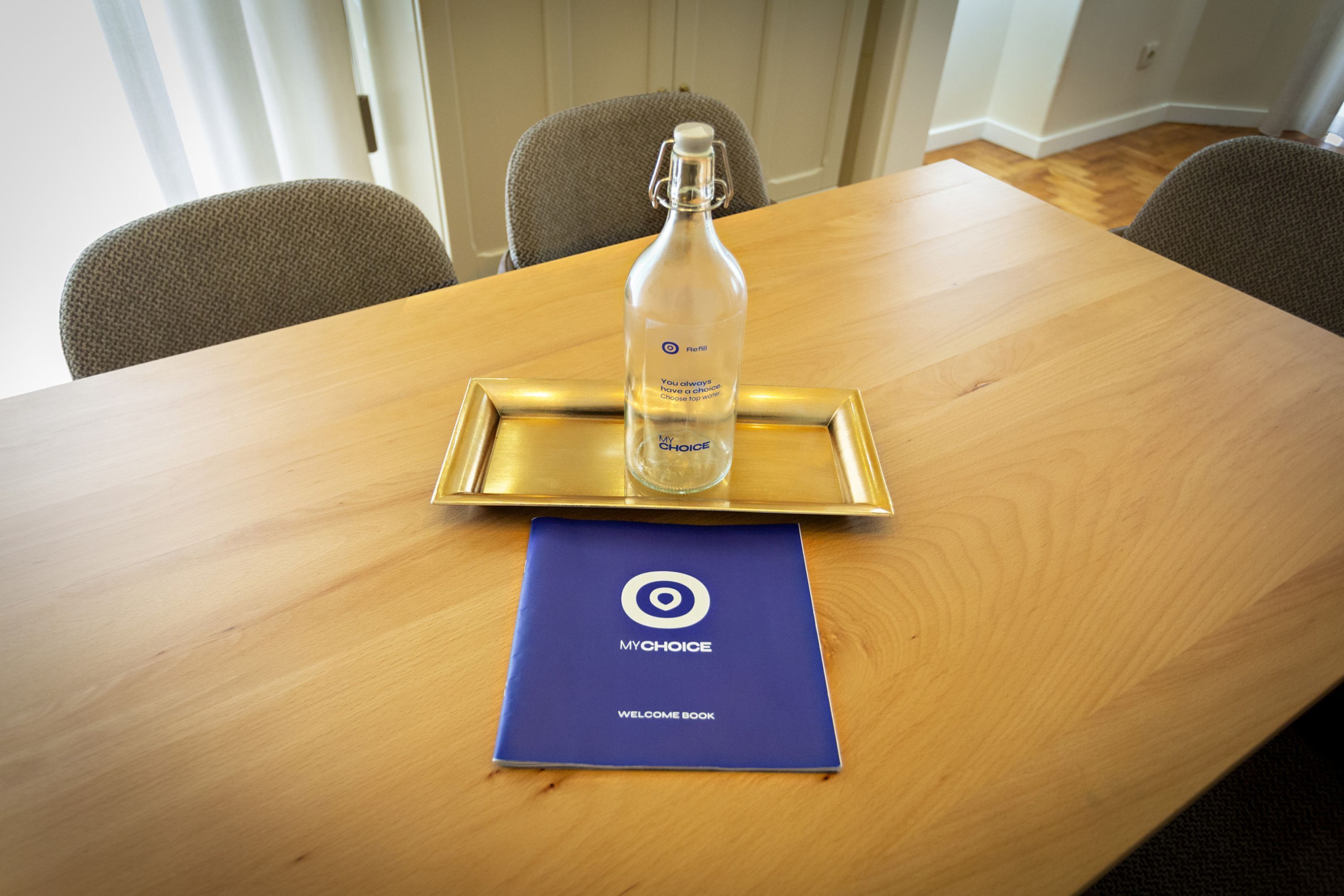 Une table en bois sur laquelle repose une bouteille en verre transparent, posée sur un plateau doré, un livre d’accueil bleu et deux chaises rembourrées.