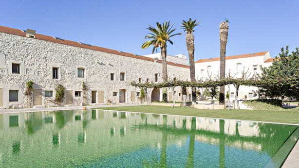 A pool area with clear water, surrounded by stone buildings with arched doorways and a palm tree. - CV BC - Convento das Bernardas (Tavira)