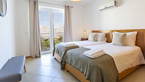 Hotellihuone, jossa on kaksi sänkyä, penkki, suuri ikkuna verhoilla, ilmastointilaite ja näkymä ulkona oleviin rakennuksiin.