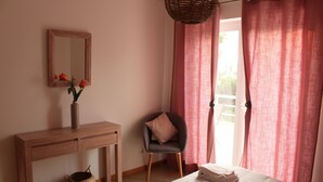 A bedroom with a wooden desk, a chair, a bed with a folded blanket, a wicker basket, and a mirror on the wall. - CG 12F - Garden - Cabanas Gardens (Tavira)
