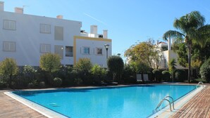 A swimming pool with a wooden deck, surrounded by greenery and modern buildings. - CG 12F - Garden - Cabanas Gardens (Tavira)