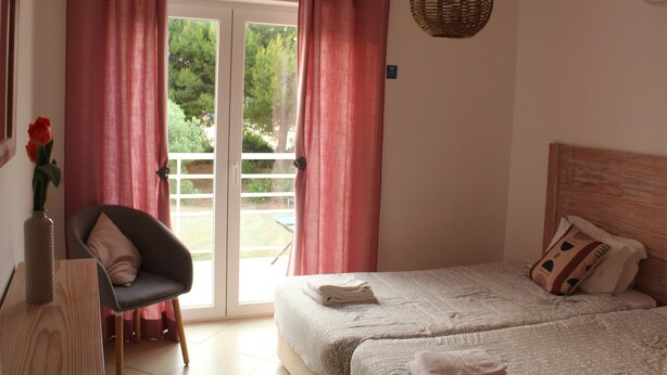 A bedroom with a bed, a chair, a vase with flowers, and a view through the door. - CG 12F - Garden - Cabanas Gardens (Tavira)