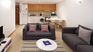 A modern living room with a grey sofa, a small coffee table, and a TV. There is a kitchen area with wooden cabinets, a washing machine, and a dining table with chairs.
