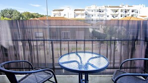 A balcony with a round table and two chairs, a metal fence, and a row of white buildings in the background.